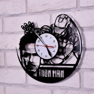 Puede incluir: Reloj de vinilo negro en forma de disco con una silueta de Iron Man. La esfera del reloj es blanca con números y manecillas negros. El reloj está montado en una pared de ladrillo blanco.