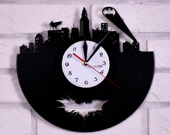 Könnte beinhalten: Schwarze Schallplattenuhr mit weißem Zifferblatt und schwarzen Zeigern. Die Uhr zeigt eine Silhouette einer Skyline mit einer darüber fliegenden Fledermaus. Die Uhr ist an einer weißen Backsteinwand montiert.