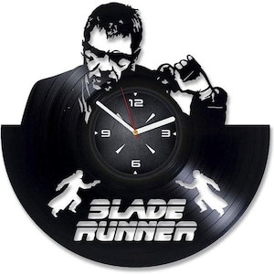 May include: Black vinyl record clock featuring a silhouette of a man and the text "BLADE RUNNER." The clock face is black with white numerals and hands. Two figures with guns are on the bottom of the clock.