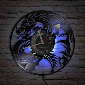 May include: A black vinyl record clock featuring a dragon design. The clock face is black with white numerals and hands. The dragon is intricately cut out, with blue backlighting illuminating the design. The clock is mounted on a brick wall.