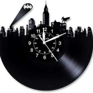 Pode incluir: Um relógio de vinil preto com uma silhueta da cidade e um logotipo do Batman. O mostrador do relógio tem números e ponteiros brancos, e o texto "Live" está impresso no mostrador.