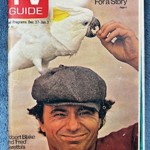 May include: A vintage TV Guide magazine cover featuring a man wearing a cap with a cockatoo perched above him. The magazine's title is in a red box, and the cover includes text about local newsmen and a story about Robert Blake.