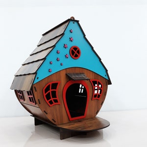 May include: A wooden house shaped like a birdhouse with a blue roof and red accents. The roof has pink stars and a red circle with a cross in the center. The house has a small porch and two windows.