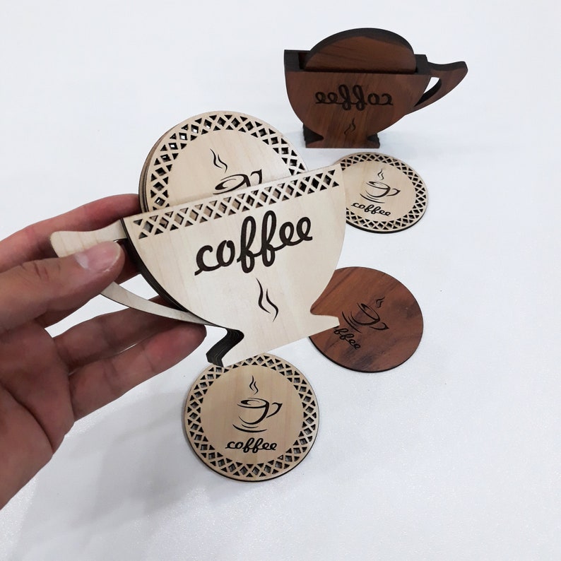 May include: Set of six wooden coasters featuring a coffee cup design. The coasters are made of light and dark wood and have a laser-etched design. The coasters are stacked in a cup shape, with the top coaster featuring the word "coffee" in a cursive font.