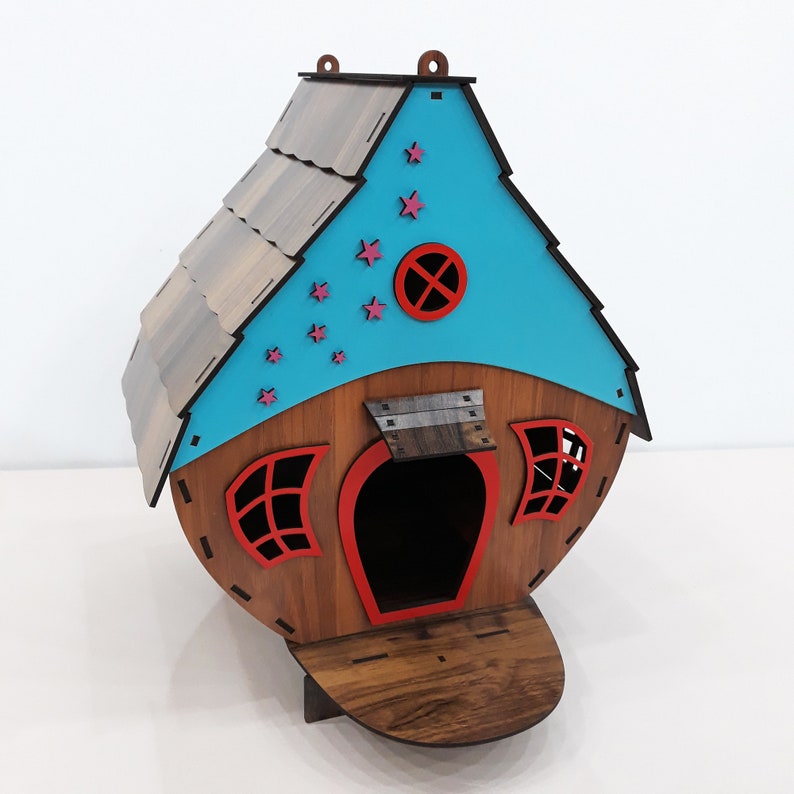 May include: A wooden birdhouse painted in shades of brown, blue, and red. The roof is brown with a blue front and a red door. The front of the house has a small red window and a red circle with a cross inside. There are pink stars painted on the blue front of the house.