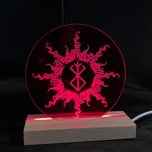 May include: A red LED night light with a circular acrylic panel featuring a stylized sun with a black and white symbol in the center. The light is on a wooden base.