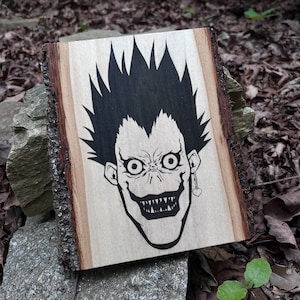 May include: A wooden plaque with a black illustration of a character's face with spiky hair, a wide grin, and large eyes. The plaque has a natural wood grain and bark edges. A heart-shaped earring hangs from one ear.