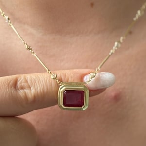 Könnte beinhalten: Eine goldene Halskette mit einem rechteckigen, in einer Zargenfassung gefassten roten Edelsteinanhänger. Die feine Kette ist mit kleinen, gleichmäßig verteilten runden Goldgliedern akzentuiert, was ein raffiniertes und strukturiertes Erscheinungsbild ergibt.