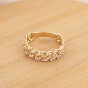 May include: A gold-colored ring with a chain-link design, encrusted with small, clear gemstones. The ring is set on a round, light-brown wooden surface, with a white fabric in the background. The ring's design is a continuous loop.