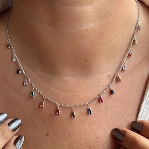 Puede incluir: Un collar de plata con una cadena delicada y múltiples colgantes en forma de lágrima de varios colores, incluyendo rojo, azul, verde y naranja. El collar se lleva alrededor del cuello.