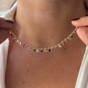 May include: A delicate rose gold necklace featuring a series of small, colorful gemstones. The gemstones are in various colors, including red, yellow, green, and blue. The necklace is held up by a person wearing a white shirt.