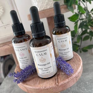 May include: Three Aromasenses essential oil bottles with black droppers are displayed on a small, round, wooden stand. The bottles are labeled "BALANCE" and "CALM", with the latter also specifying "Orange Geranium Lavender" and "Massage Oil". Lavender sprigs are placed on the stand.