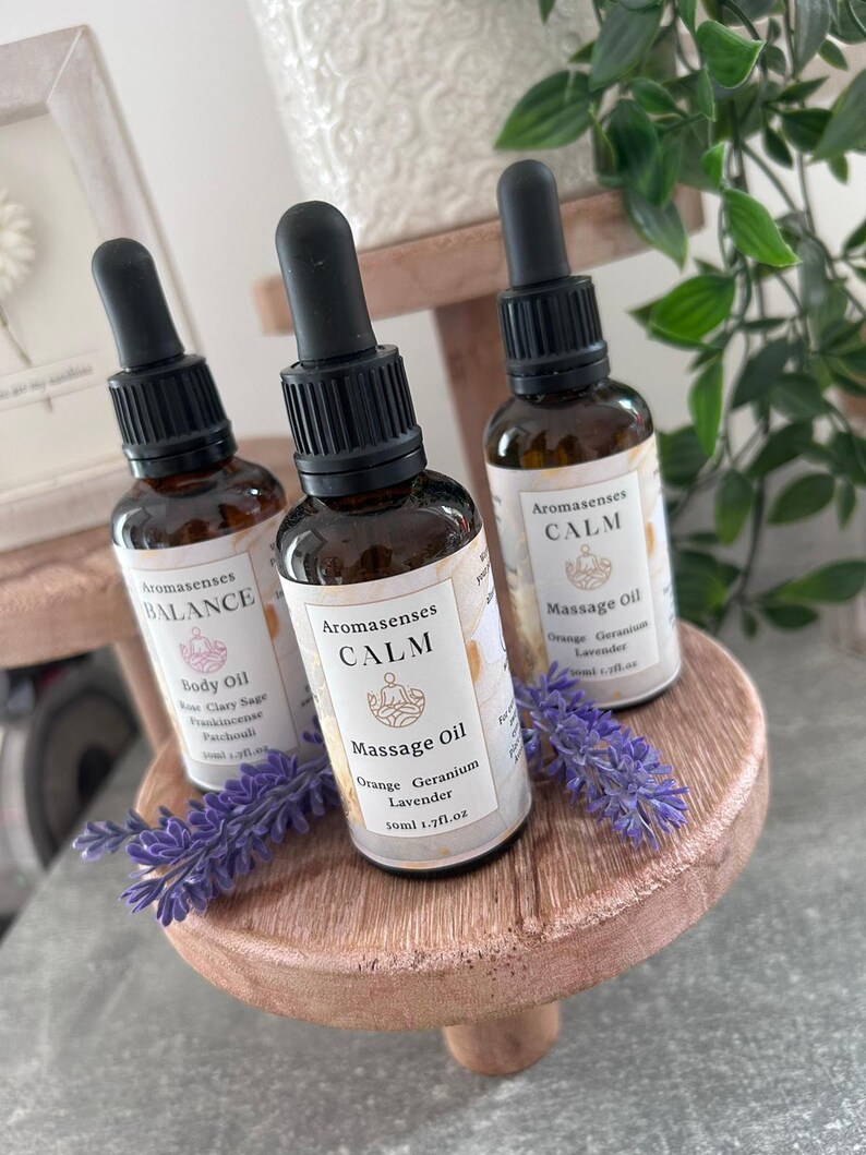 May include: Three Aromasenses essential oil bottles with black droppers are displayed on a small, round, wooden stand. The bottles are labeled "BALANCE" and "CALM", with the latter also specifying "Orange Geranium Lavender" and "Massage Oil". Lavender sprigs are placed on the stand.