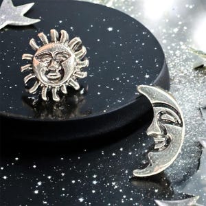 May include: A silver sun and moon charm set with a black background. The sun and moon have faces and are in a crescent shape.