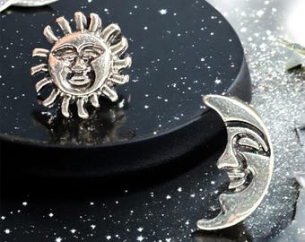 May include: A silver sun and moon charm set with a black background. The sun and moon have faces and are in a crescent shape.