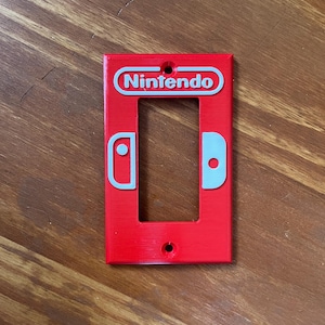 May include: A red Nintendo light switch cover with the Nintendo logo and controller icons in light blue. The cover has a rectangular shape with a single opening for the switch and two screw holes.