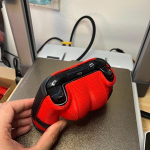 May include: A red and black gaming controller with a textured grip. The controller is on a gray surface.