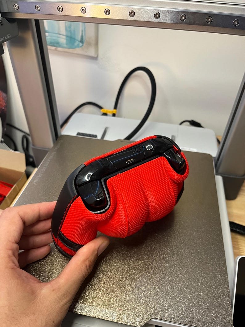 May include: A red and black gaming controller with a textured grip. The controller is on a gray surface.