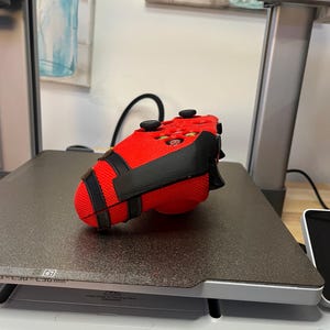May include: A red and black 3D printed video game controller. The controller is sitting on a black surface.