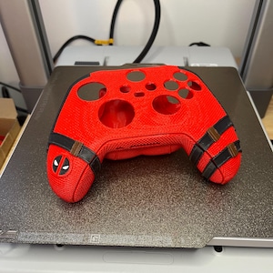 May include: A red 3D printed video game controller shell with black accents and a Deadpool logo. The shell is designed to fit a standard Xbox controller.