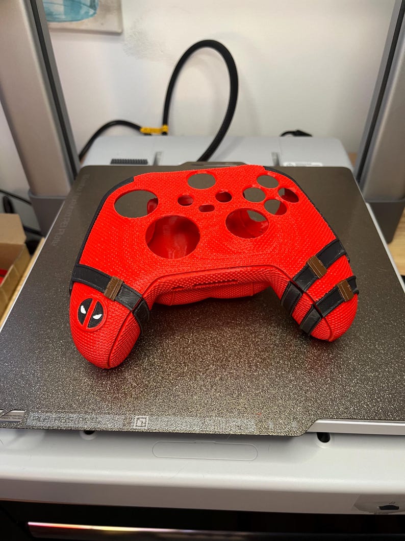 May include: A red 3D printed video game controller shell with black accents and a Deadpool logo. The shell is designed to fit a standard Xbox controller.