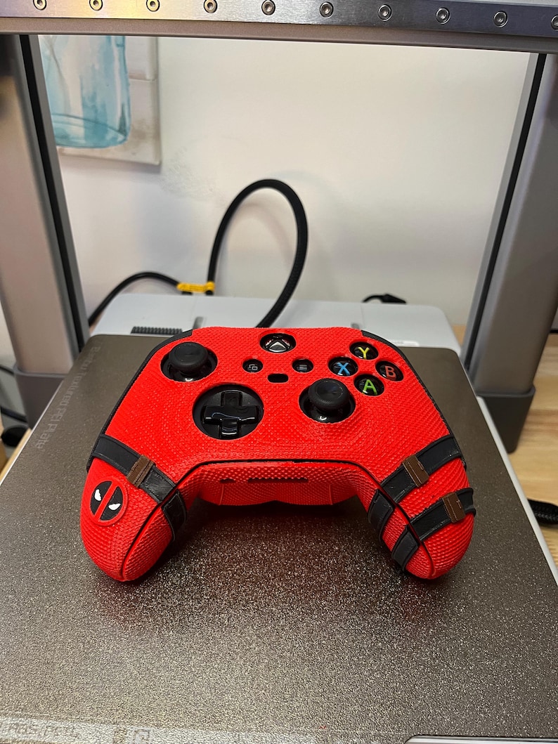 May include: Red and black video game controller with a Deadpool design. The controller has a textured surface and black straps with a Deadpool logo.