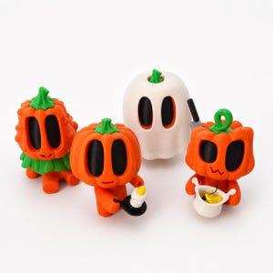 May include: Four Halloween-themed figurines. Two orange pumpkin figures with green stems and black oval eyes. A white ghost figure with black oval eyes and a green stem. One pumpkin figure holds a small white bowl. The other holds a candle.
