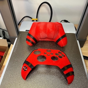 May include: Red 3D printed video game controller shell with black straps and a Deadpool logo. The shell is designed to fit a standard Xbox controller.