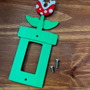 May include: A green light switch cover featuring a red and white flower design. The flower has white spots and a red mouth. Two screws are included. The design is inspired by a popular video game.
