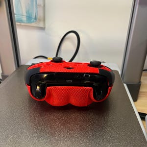 May include: A red and black video game controller with a textured surface. The controller is lying on a gray surface.