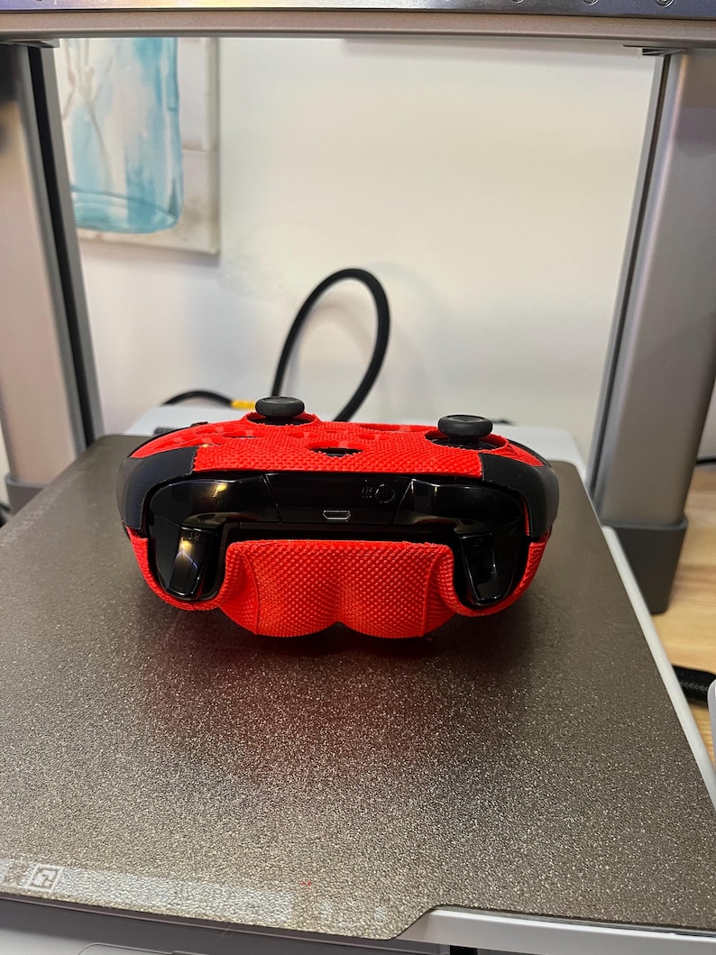 May include: A red and black video game controller with a textured surface. The controller is lying on a gray surface.