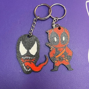 Op de afbeelding: Een sleutelhanger met twee cartoonfiguren. De ene sleutelhanger heeft een zwart-wit Venom-figuur met een rode tong. De andere sleutelhanger heeft een rood-zwart Deadpool-figuur.