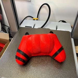 May include: A red 3D printed video game controller with black accents. The controller is sitting on a gray textured surface.