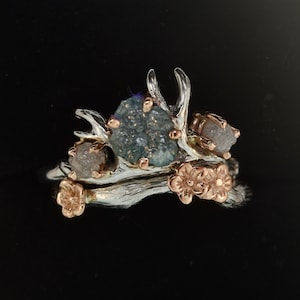 May include: A ring featuring a textured silver band with rose gold flower accents and branch-like prongs. The setting holds a central blue druzy stone flanked by two smaller, rough, light-colored stones, all secured by rose gold metal prongs.