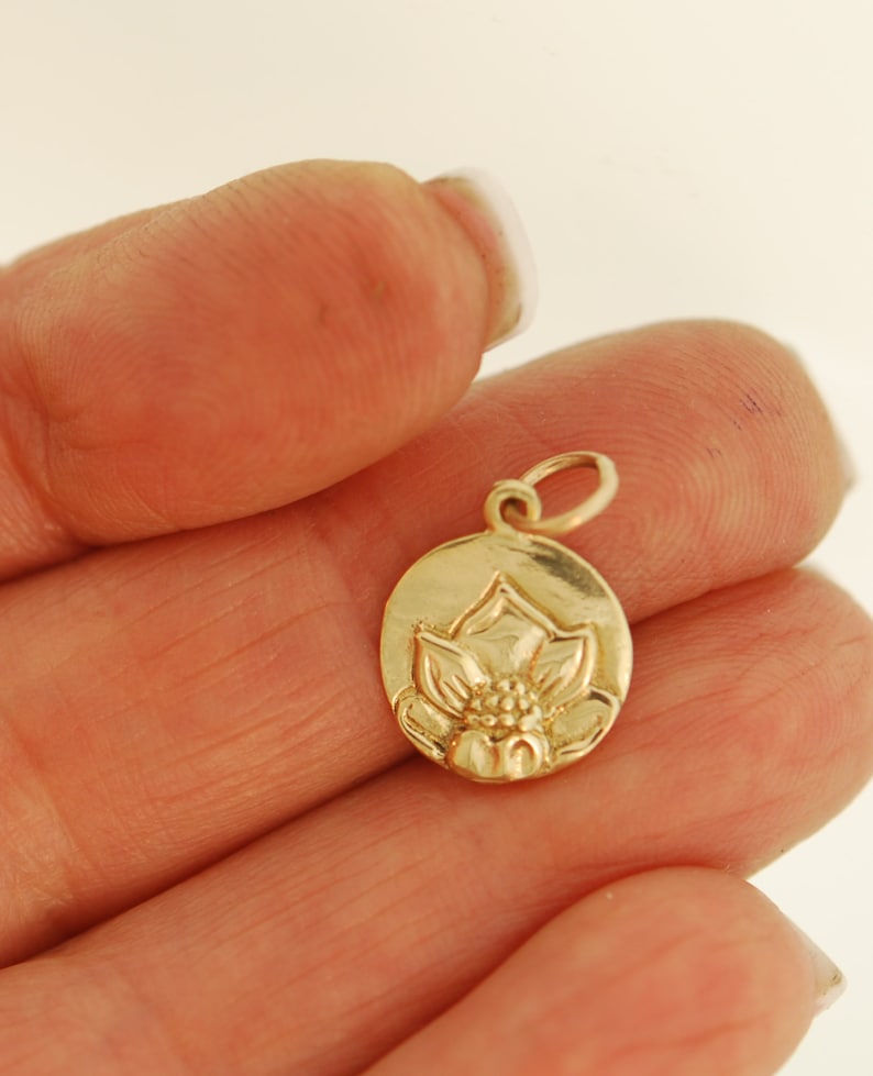 May include: A gold-colored charm featuring a detailed lotus flower design. The charm is oval-shaped and has a loop at the top for attaching to a necklace or bracelet.
