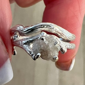 May include: A silver ring set featuring a textured band resembling a tree branch and a second band holding a rough, uncut, translucent white gemstone held in place by four prongs. The metal has a polished, reflective finish with organic details.