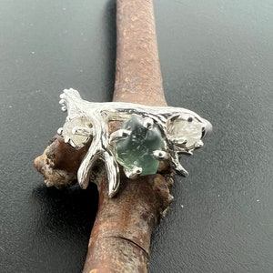 May include: A silver ring with a branch design featuring a green gemstone and two clear stones.