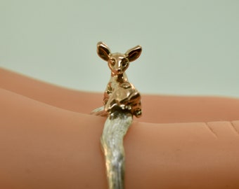 May include: A gold-toned ring featuring a detailed deer figurine. The deer is sitting on a silver band that wraps around the finger.
