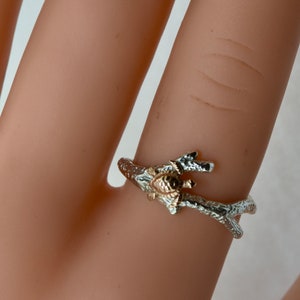 May include: A silver ring with a gold turtle design on a branch. The ring is on a finger.