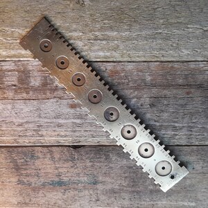 May include: A metal ring sizer tool with numbered markings and circular cutouts for sizing rings. The tool is silver in colour and has a worn, vintage look.