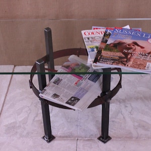 May include: A glass-topped coffee table with a metal frame and a brown leather strap. The table has three legs and is decorated with magazines and a newspaper.