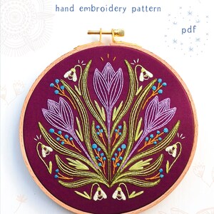 May include: Embroidered hoop art featuring a floral design with purple crocuses, green leaves, and blue berries. The hoop is on a white background with the text "EARLY BLOOMER" and "COZYBLUE handmade" in black.