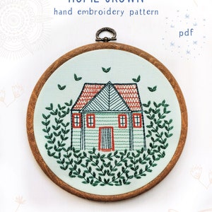 May include: Embroidered hoop art featuring a green house with a red roof and a white door. The house is surrounded by green leaves and the hoop is made of wood.