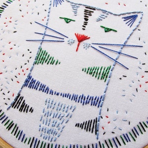 May include: Embroidered cat design on a white fabric stretched over a wooden hoop. The cat is blue with green eyes and a red nose. The background is white with black, red, blue, and green embroidery.