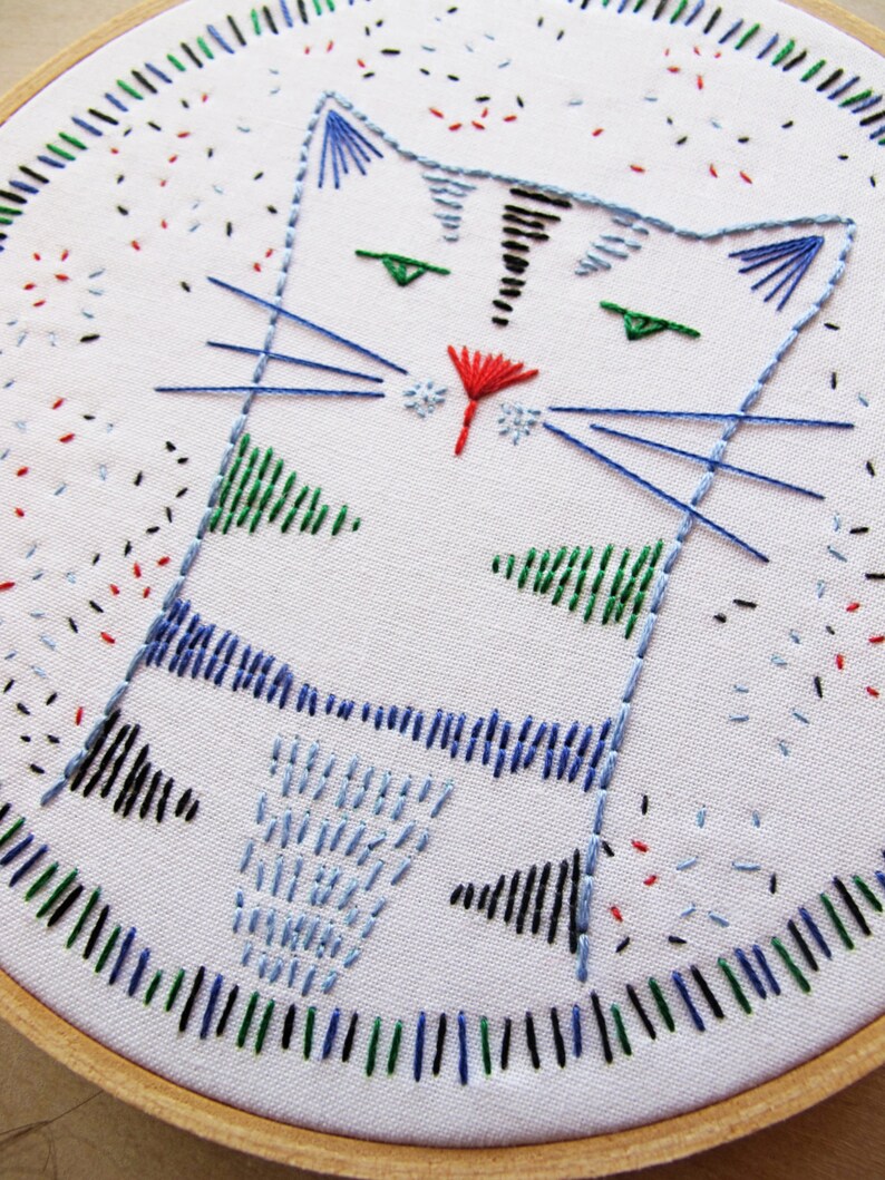 May include: Embroidered cat design on a white fabric stretched over a wooden hoop. The cat is blue with green eyes and a red nose. The background is white with black, red, blue, and green embroidery.