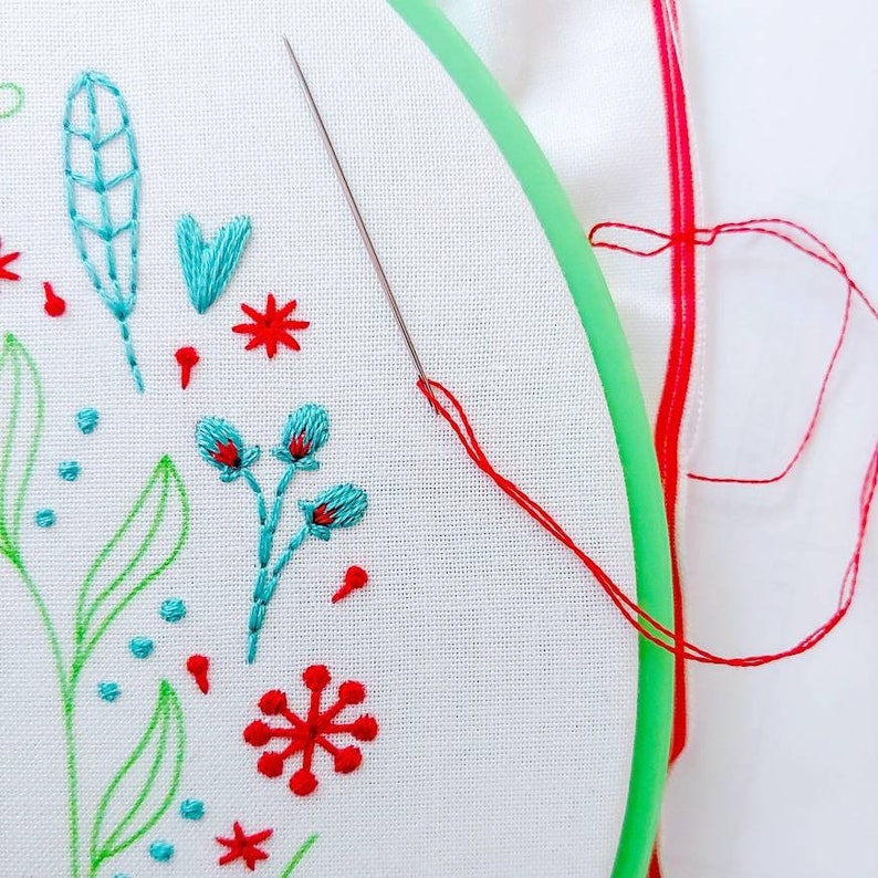 May include: A close-up of a white fabric with embroidery of green leaves, blue flowers, and red flowers. A needle and red thread are visible in the image.