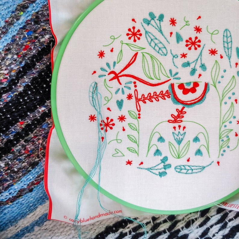 May include: A white embroidery hoop with a green and red floral design. The design includes flowers, leaves, and stems. The hoop is on a blue and black patterned fabric.