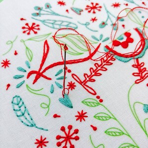 May include: Close-up of a white fabric with red and green embroidery. The embroidery features a variety of floral designs, including leaves, flowers, and stems. A needle and thread are visible in the center of the image.