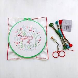 May include: A green embroidery hoop with a white fabric inside. The fabric has a red and green embroidered design of a Dala horse with flowers. The hoop is on a white surface with red, blue, and green embroidery floss, a small glass bottle, and a pair of red embroidery scissors.