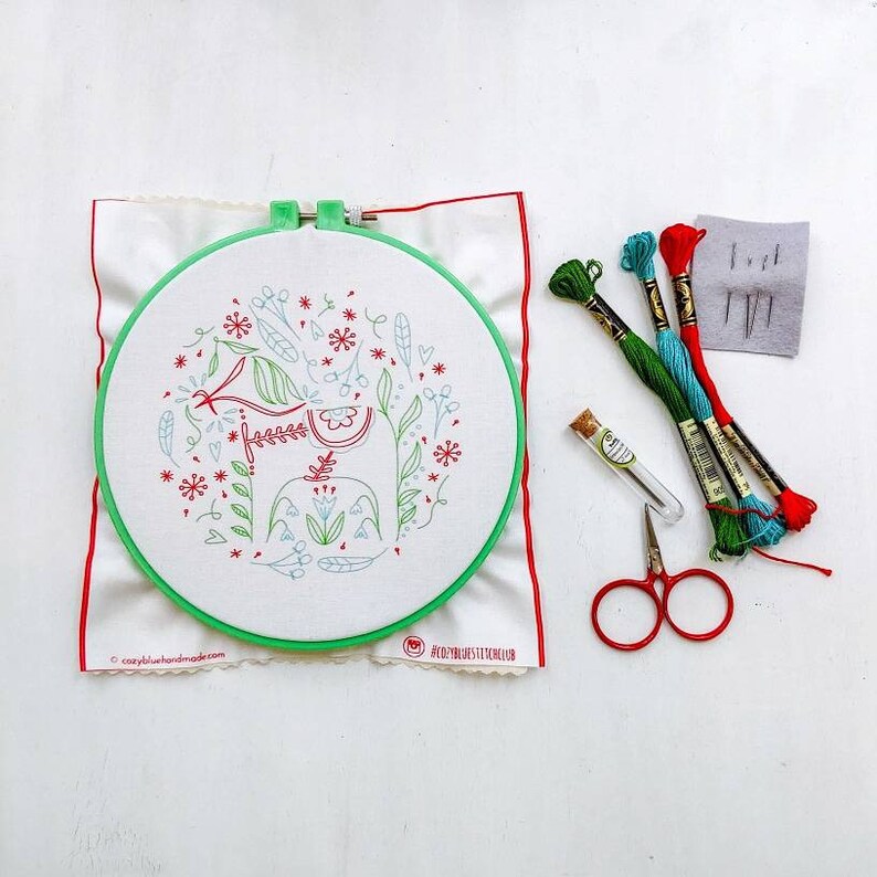 May include: A green embroidery hoop with a white fabric inside. The fabric has a red and green embroidered design of a Dala horse with flowers. The hoop is on a white surface with red, blue, and green embroidery floss, a small glass bottle, and a pair of red embroidery scissors.